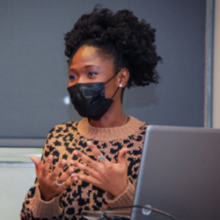 A woman in a mask stands to give a presentation.