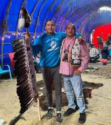 Jordan Williams White Eye and another man stand with an eagle staff inside a longhouse-style camp.