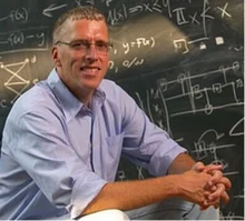 Dr. Cliff Burgess sits in front of a blackboard covered in formulae.
