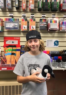 A boy wearing a University of Waterloo ball cap holds a plush goose in the Book Store while standing in front of school supplies.