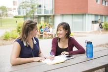 Two students sit outside talking, one is taking notes.