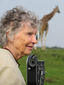 Anne Dagg behind a camera with a giraffe in the background.