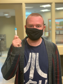 A masked man holds a sealed tube containing a COVID swab.