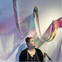 An artwork entitled &quot;Edge of Sky&quot; featuring a woman looking up and smiling at a multicoloured expanse overhead.