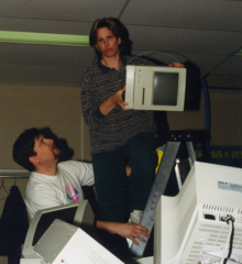 Mary Burden stands astride a ladder holding a Macintosh computer with a pile of CRT monitors below her.