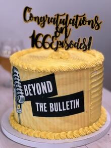 A tall cake with yellow frosting with the Beyond the Bulletin logo silkscreened on the side and a topper that says &quot;Congratulations on 166 episodes!&quot;