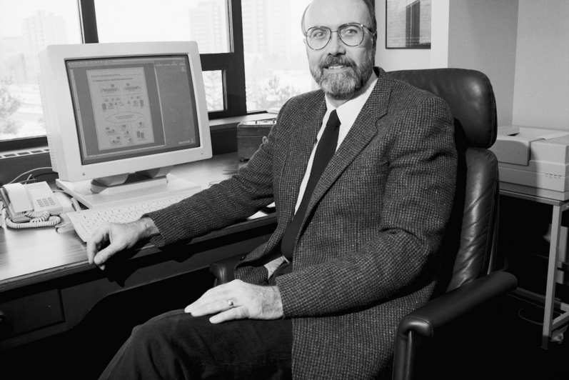Dr. Paul Guild sits at a desk in his office with a PC monitor close by.