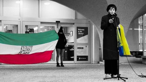 A speaker at a campus rally holds a Ukrainian flag while other students hold an Iranian flag in the background.