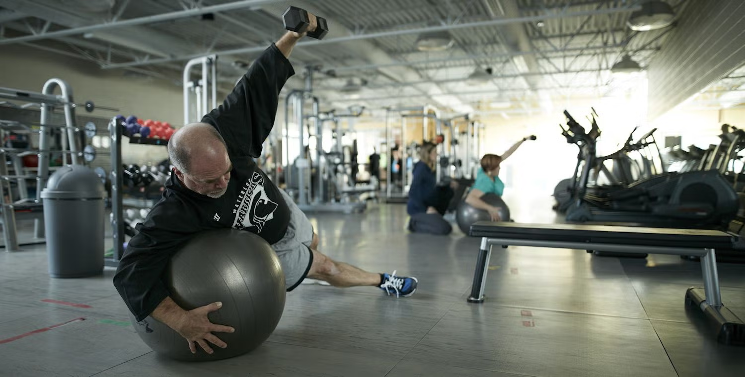 A man works out on a yoga ball while holding a dumbbell in a gym environment.