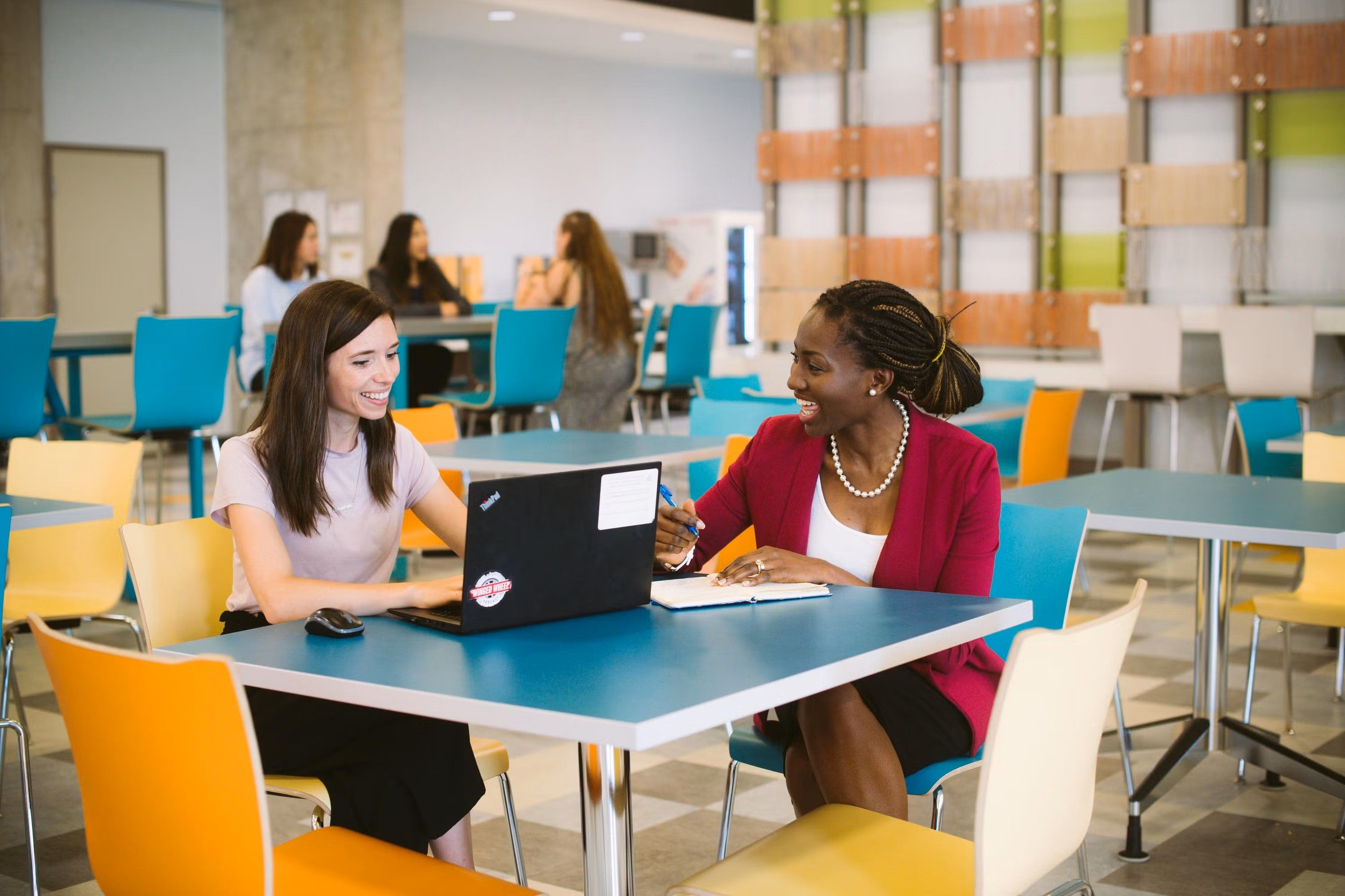 Two women sit at a table having a discussion using a laptop and notebook