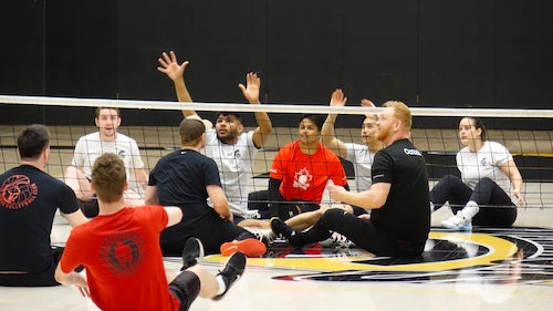 Athletes sitting on a floor with a volleyball net between them as part of a sitting volleyball game.