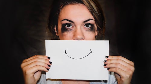 A woman with mascara rings around her eyes holds up a card over her mouth with a smile on it.