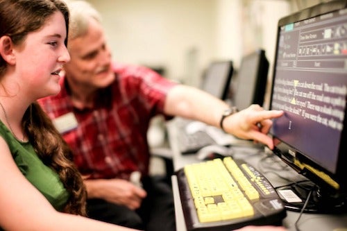 An instructor assists a patient using a computer with vision aids on the screen and keyboard.