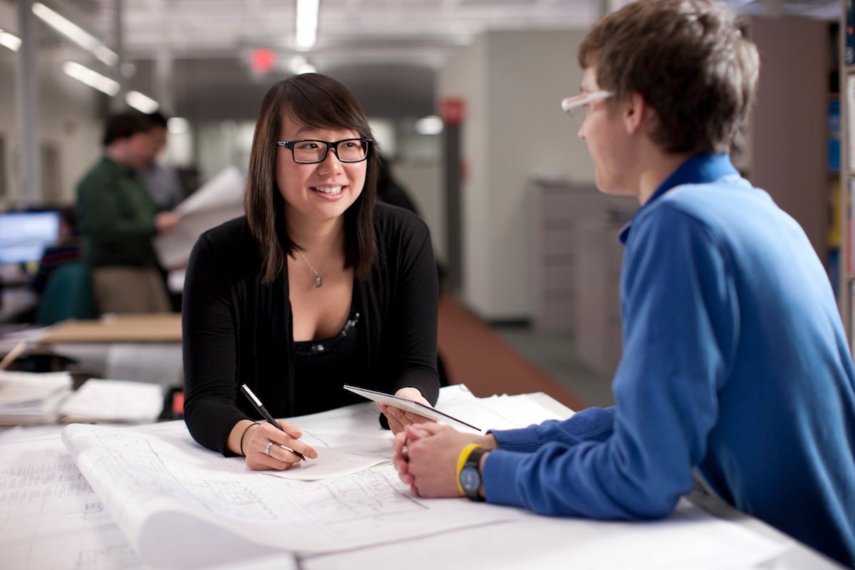 A woman and man sit together at a table, smiling while they are chatting about work set out in front of them. 