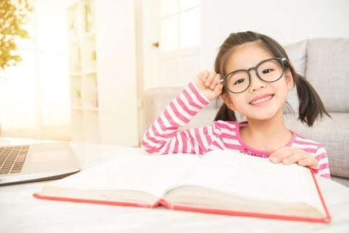 A child adjusts her glasses as she reads a book and smiles.
