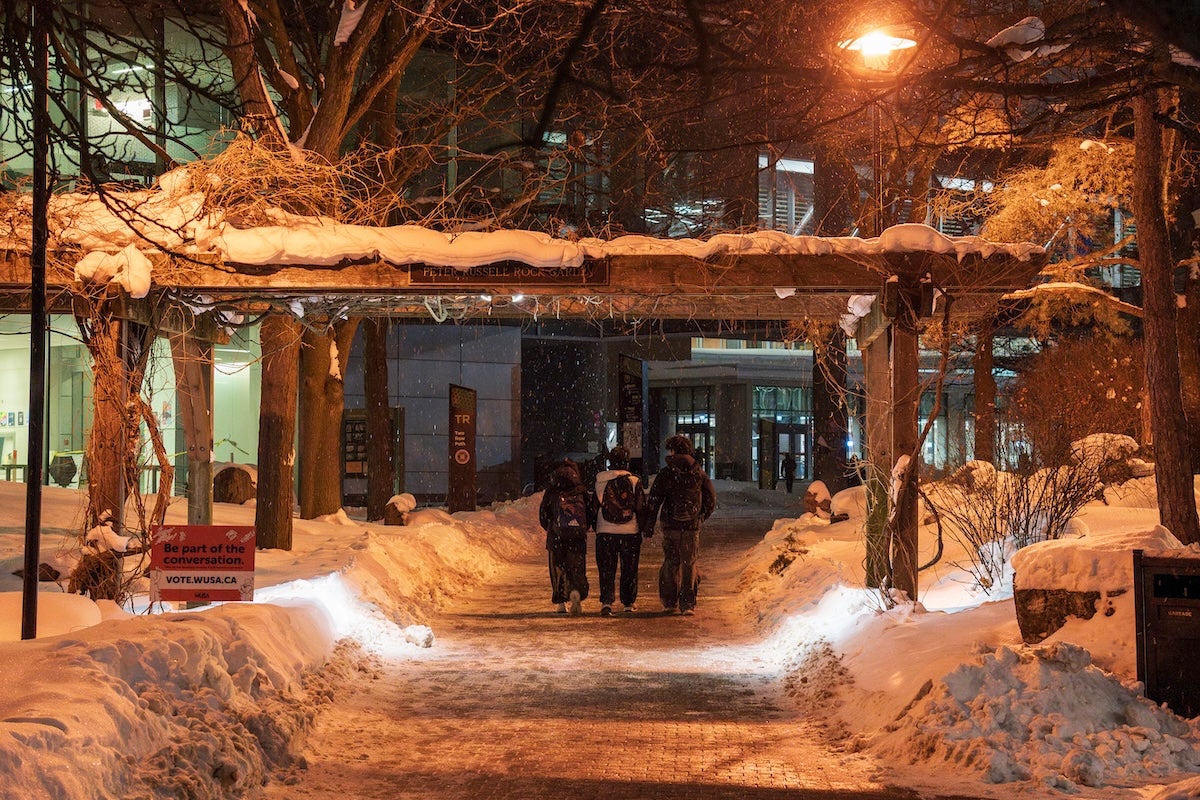 Three students walk between the snow-laden trellis of the Peter Russell Rock Garden at night.