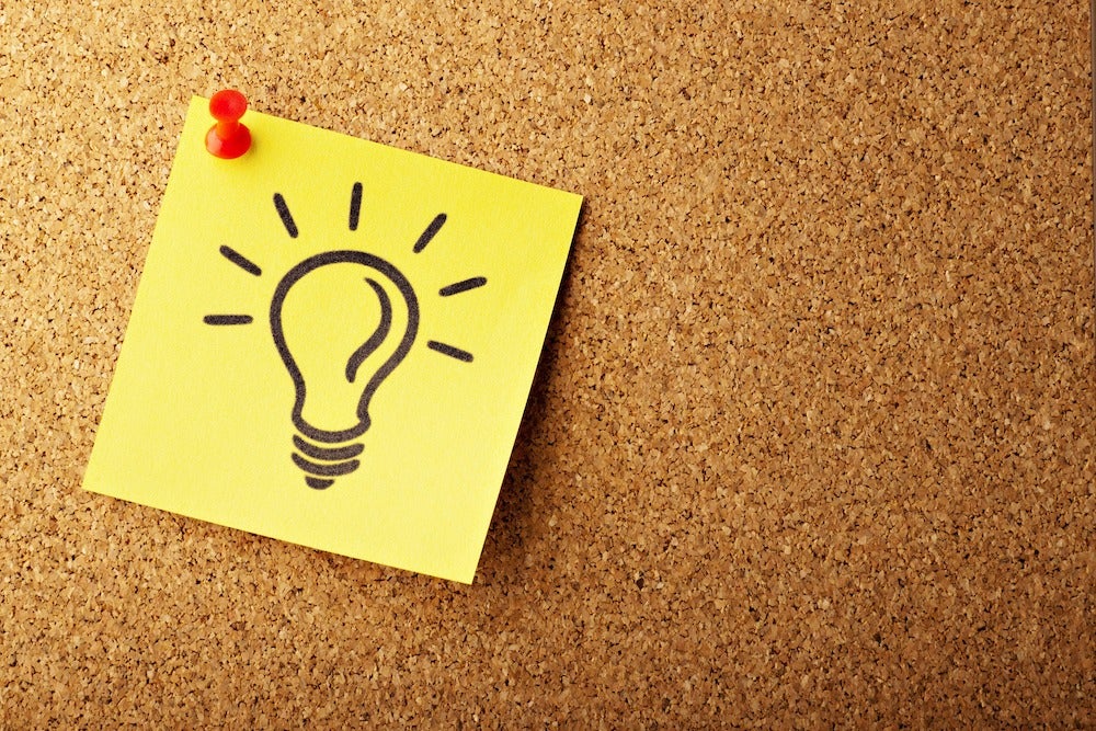 A sticky note with an illuminated light bulb is tacked to a corkboard.