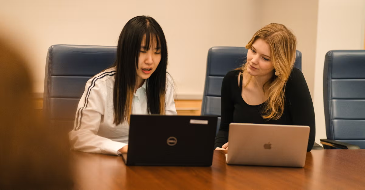 Two women sit at a table with their laptops.