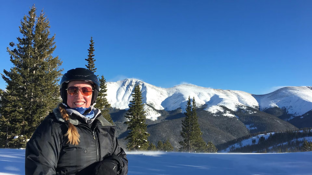 Kristine Dalton wears a helmet and sunglasses as she stands in front of a snow-capped mountain range.