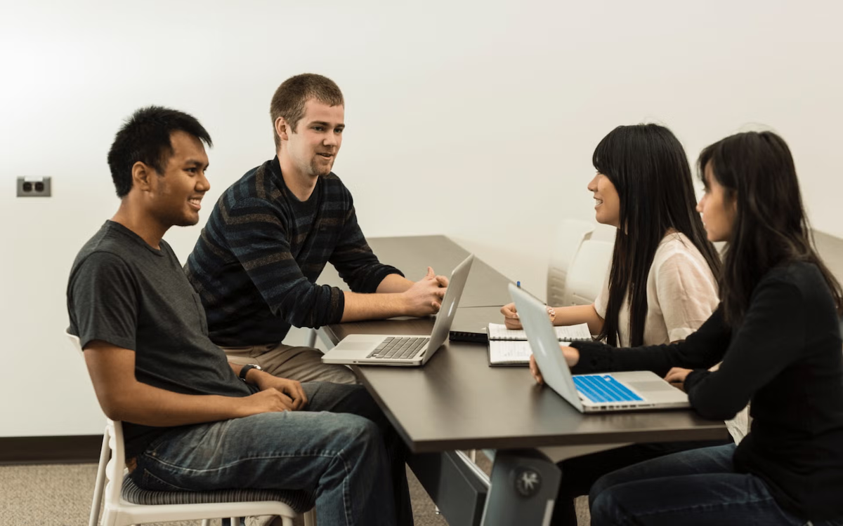 Four students converse at a table with their laptops open.