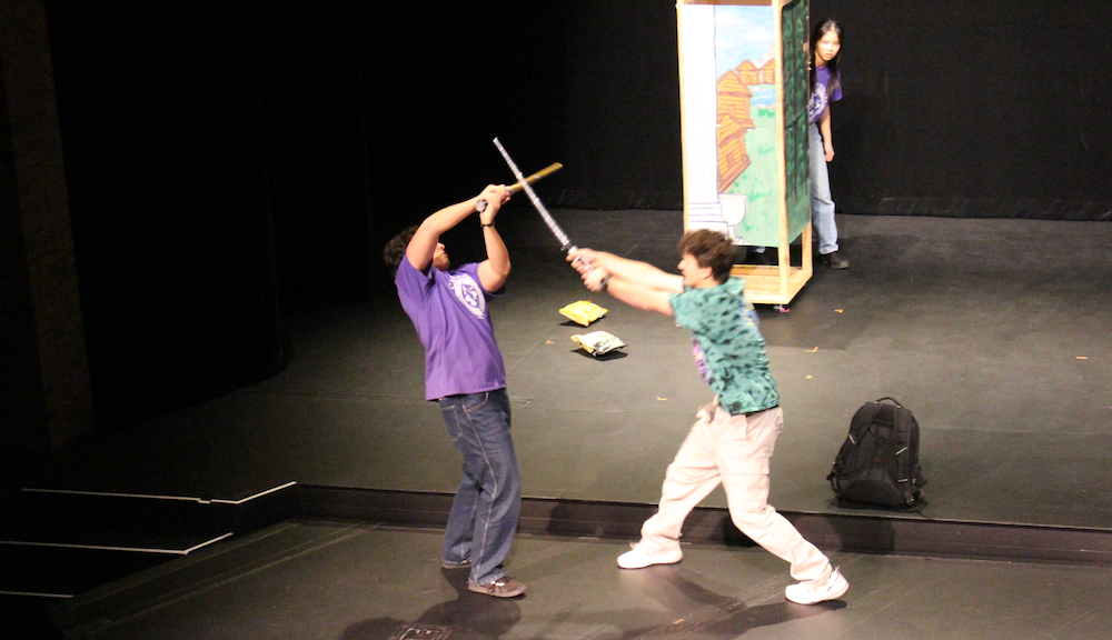 Two stage actors fight with swords while an actress peeks from behind a pylon.