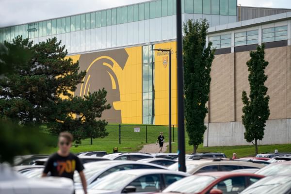 A man walks through a parking lot with the Field House in the background.
