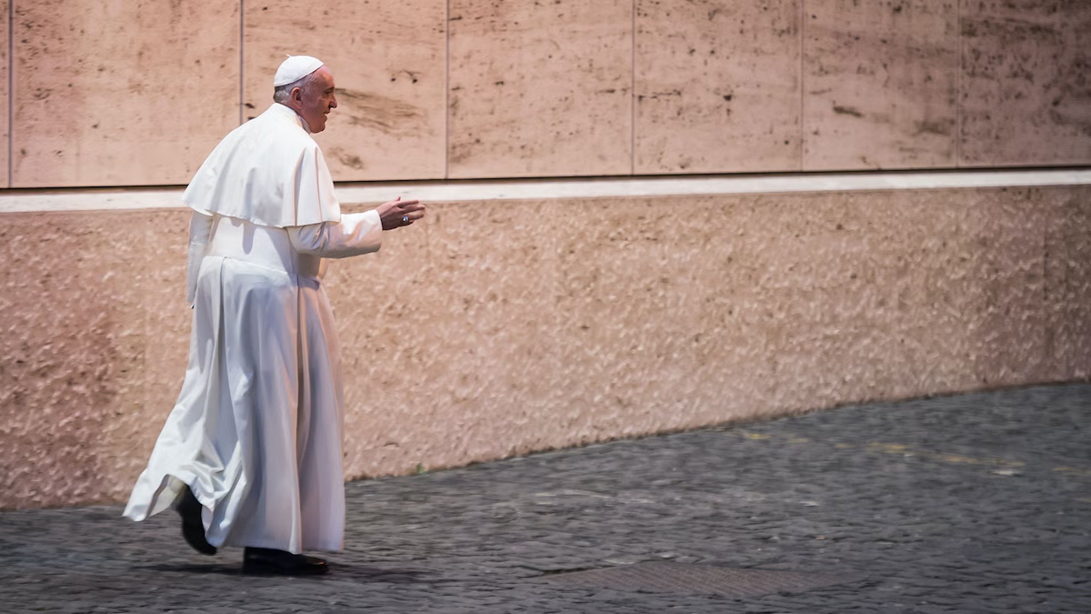 Pope Francis, wearing vestments, walks along a wall.