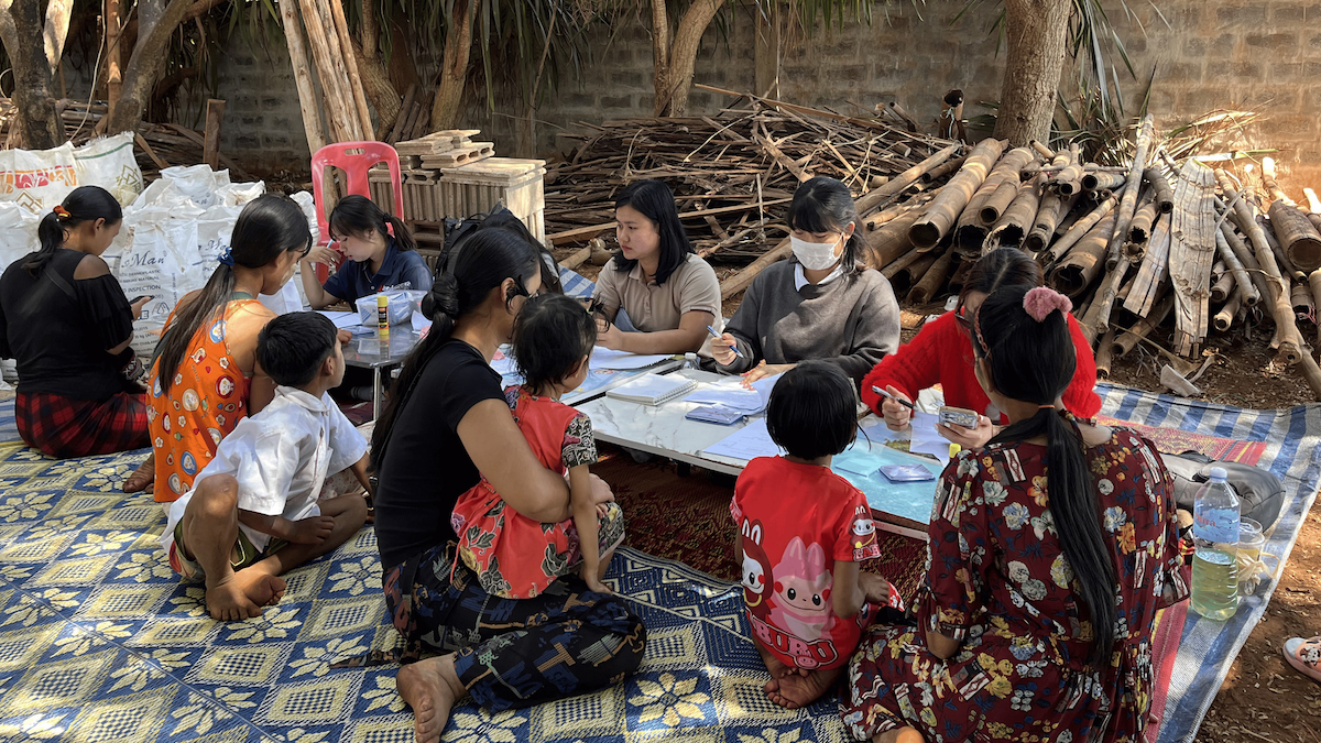 Refugees work on schoolwork at a long table surrounded by bags of rice.