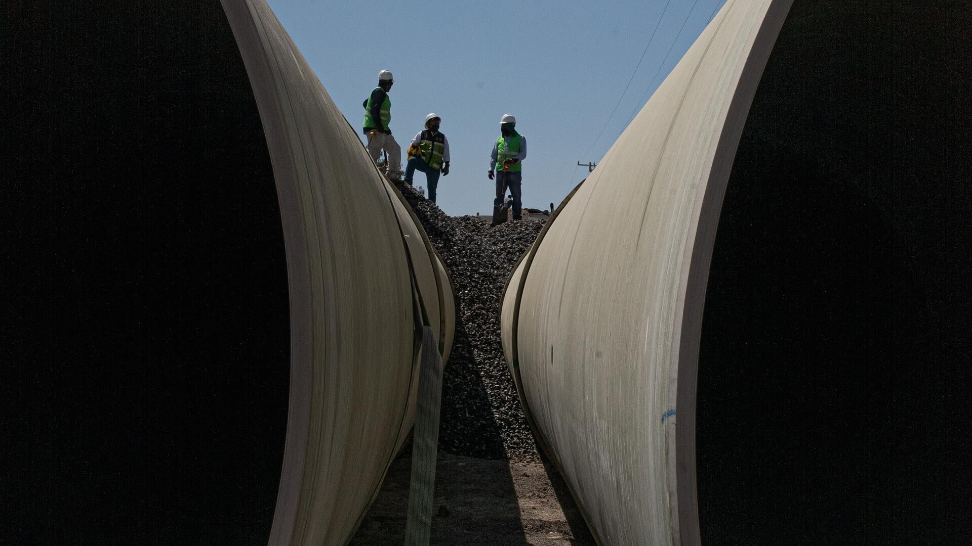 Workers in hard hats stand above two half-buried pipes in the ground