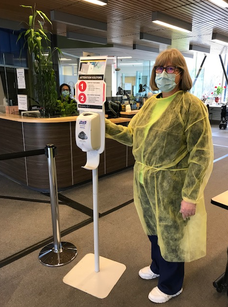 A Health Services staffer in PPE gear stands next to a sanitizer station.