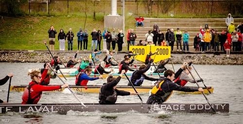 Teams rowing concrete canoes cross the finish line at a canoe competition.