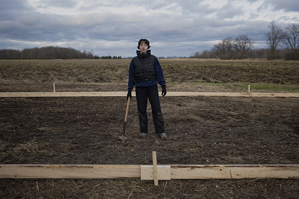 Artist Elise Popa stands in a plot of land with a gardening implement.