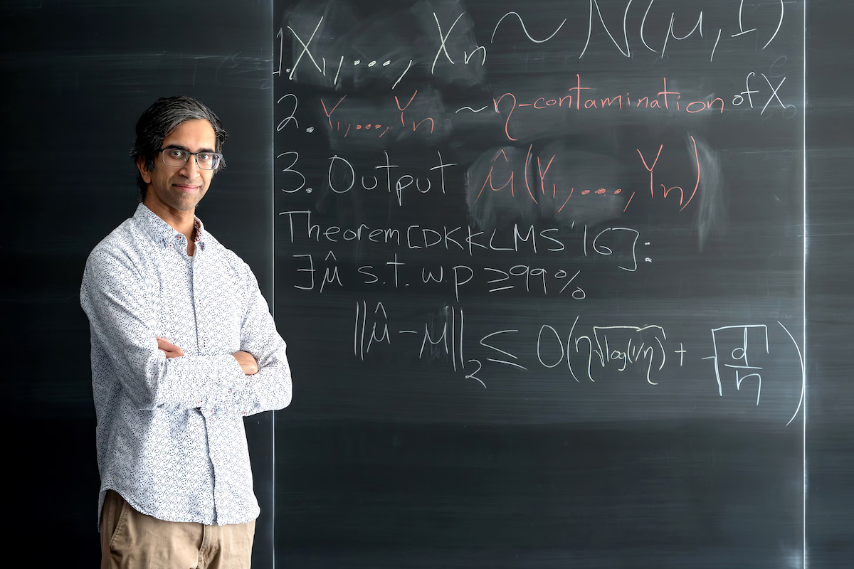 Professor Gautam Kamath stands in front of a blackboard with equations on it.