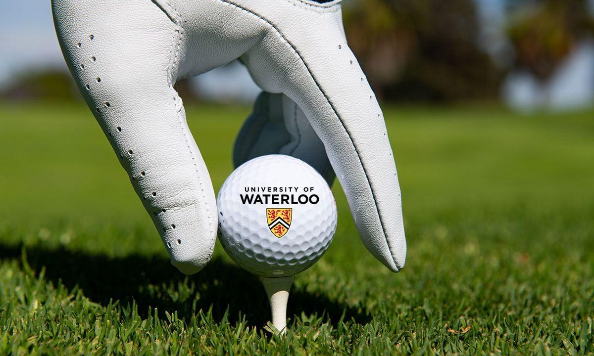 A hand wearing golfer's gloves places a University of Waterloo golf ball on a tee.