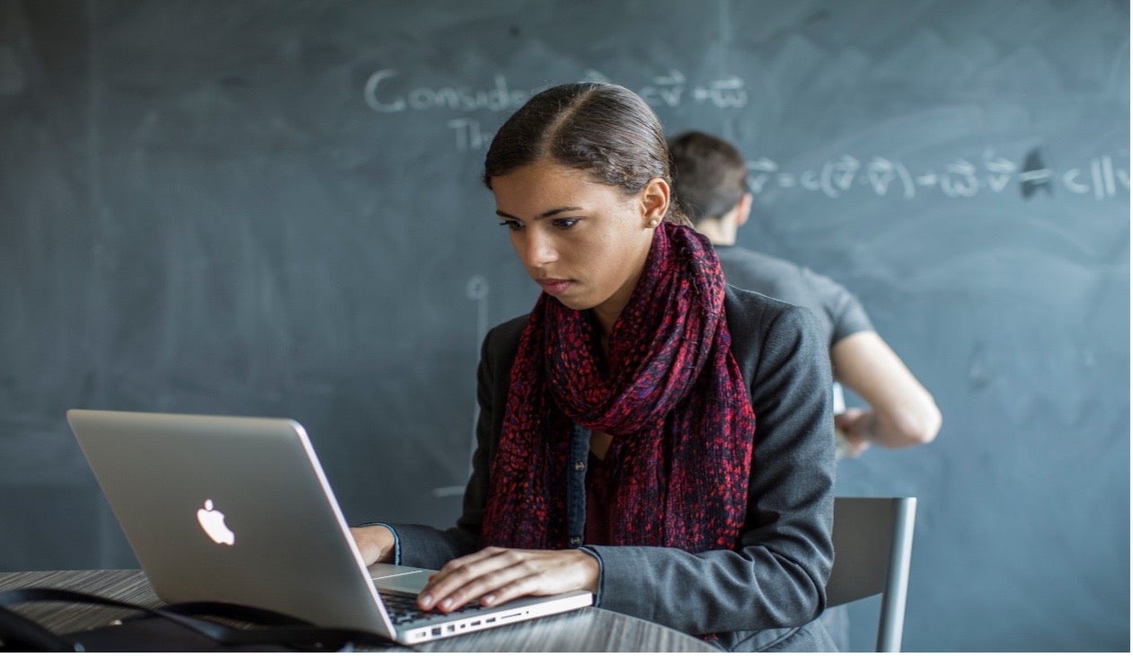 A student works on a laptop while another student behind her writes on a blackboard.