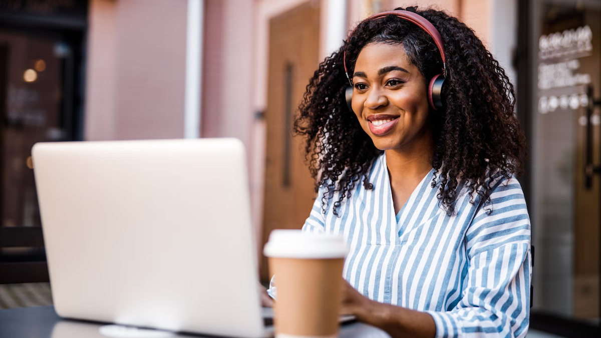 A woman wearing headphones works on a laptop with a cup of coffee within reach.