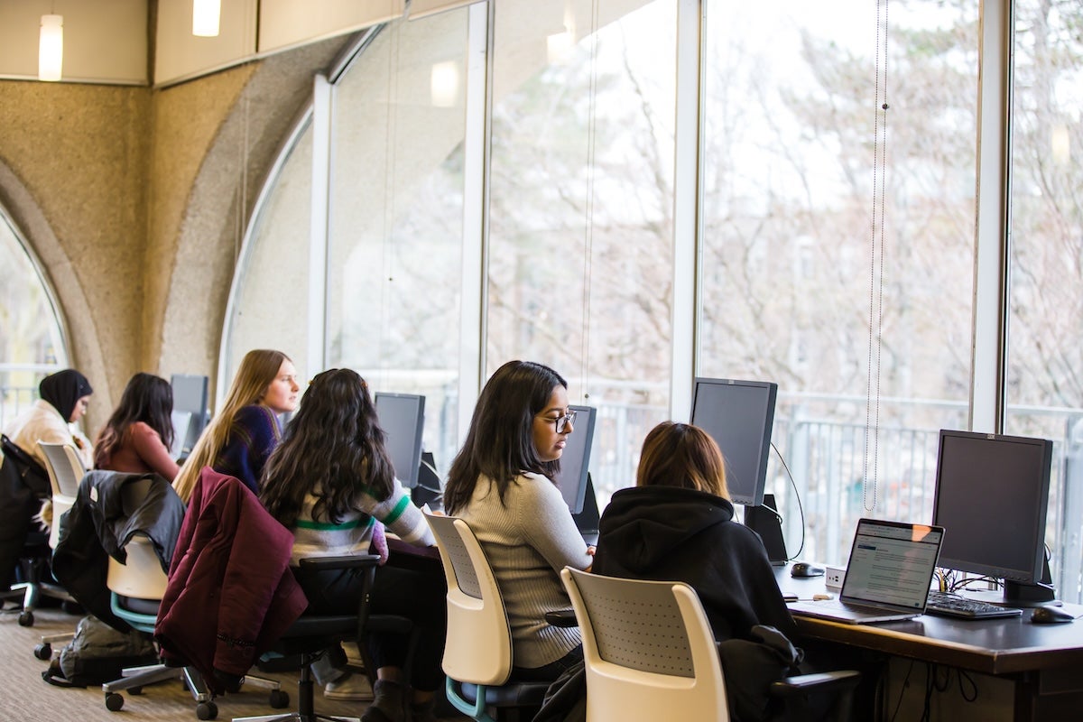 Students seated at a row of desktop computers in a bright, modern study space with large curved windows.