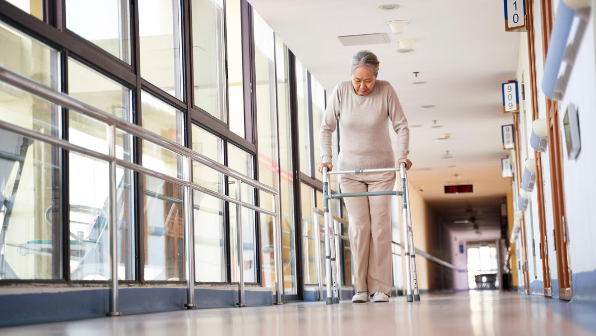 A woman walks with the support of a walker in a brightly lit hallway.