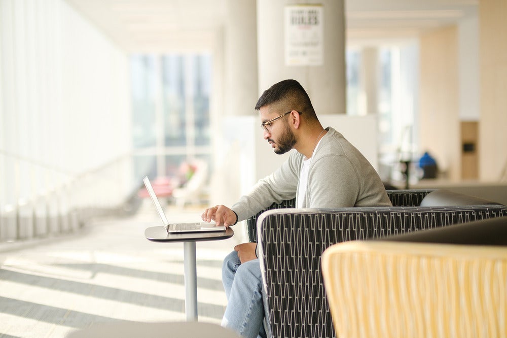 Person seated in a modern lounge area, working on a laptop placed on a small round table, with soft daylight and blurred seating and windows in the background.