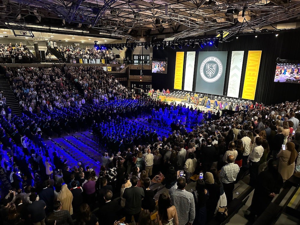 The audience stands during the Convocation ceremony in the PAC with the stage in the background.