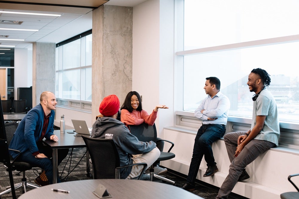 Five MBET (Master of Business, Entrepreneurship and Technology) graduate students sit around a table and on a window ledge in a classroom, actively collaborating on a business venture project.