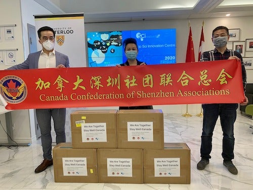 Representatives from the University of Waterloo and Sci International stand with boxes of facemasks and a supportive banner.