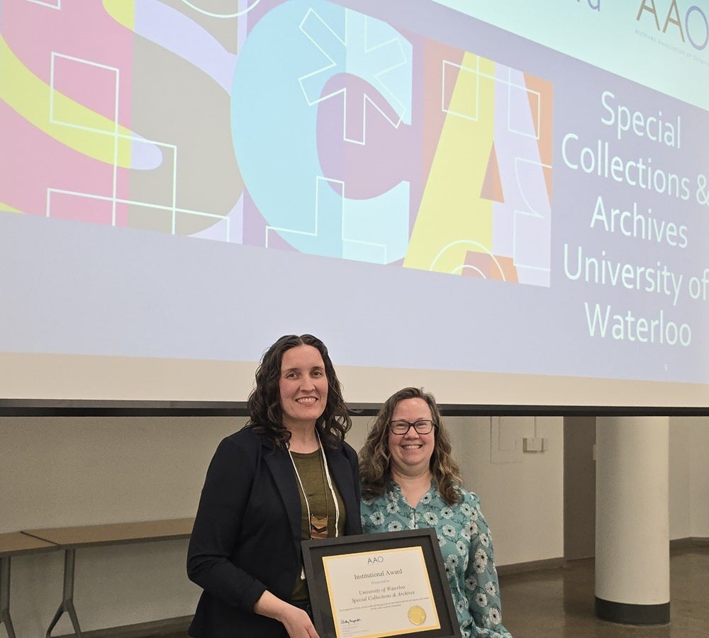Two people stand together at the front of a room holding a framed certificate. Behind them, a large projected slide displays a colorful abstract design.