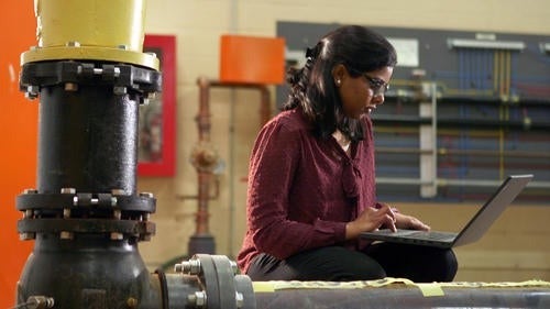 Pampa Dey works on a laptop with a large pipe junction in the foreground.