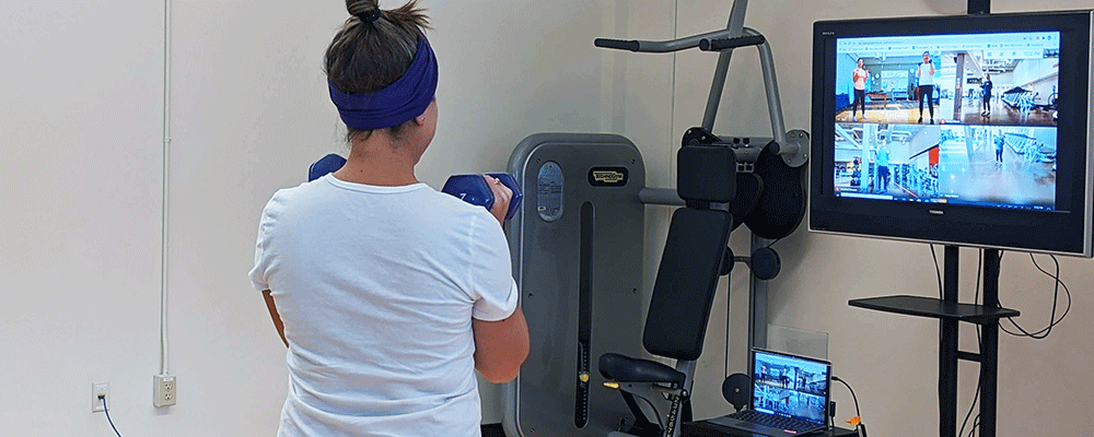 A woman lifts dumbbells in a gym in front of a video screen.