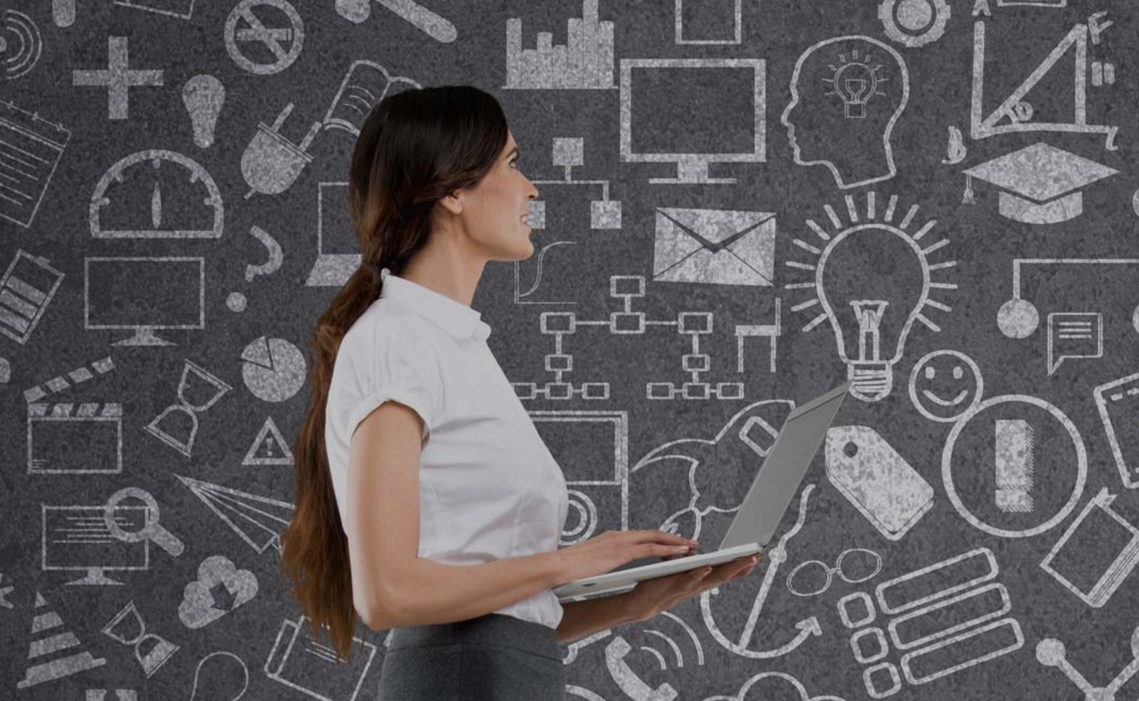 A woman with a laptop stands before a chalkboard covered in symbols.