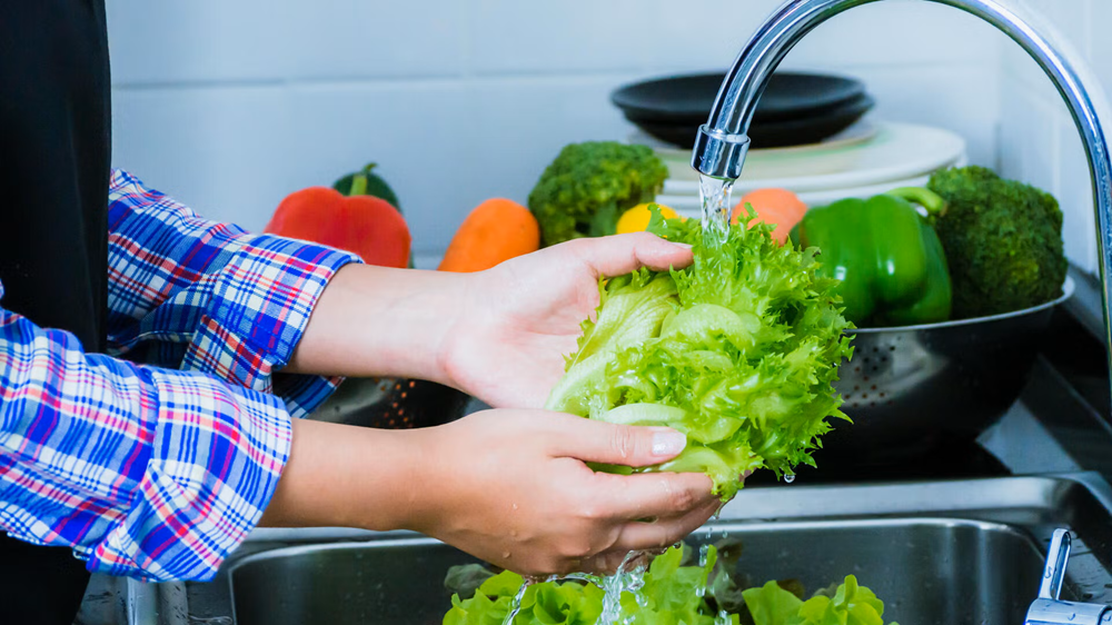 A person holds lettuce under a running tap with other vegetables on the counter in the background.