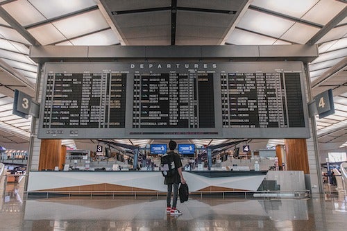 A woman stands before a departures listing at an airport.