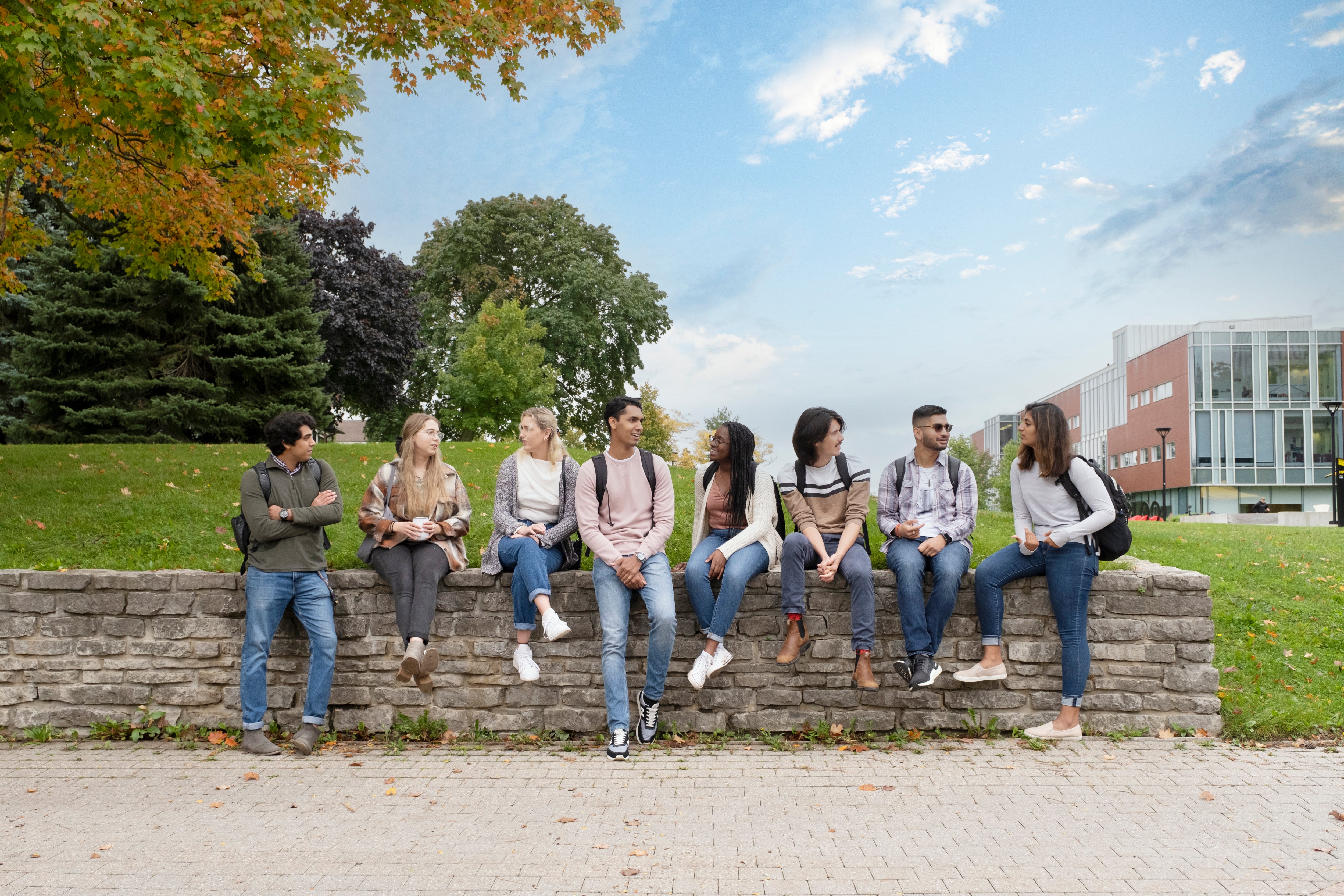 Students sit and chat along a stone wall on the University of Waterloo campus, with trees and campus buildings in the background.