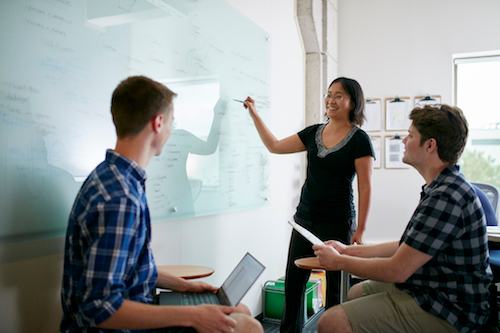 A person writes on a whiteboard while two other people look on.